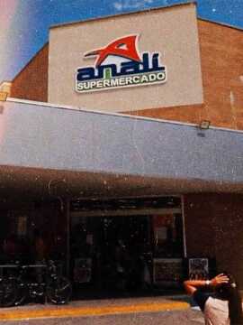 Foto de uma entrada frontal do mercado Anali. Acima da entrada a um logo com a letra 'A' destacada na cor vermelha com o nome completo logo abaixo na cor azul.