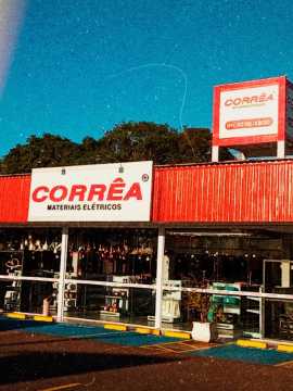 Foto frontal da Corrêa Materiais Elétricos. Na parte superior do prédio uma fachada vermelha com placa uma placa branca com Corrêa em vermelho.