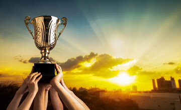 Fundo com um por do sol e, ao lado esquerda, mãos de pessoas levantando um troféu.